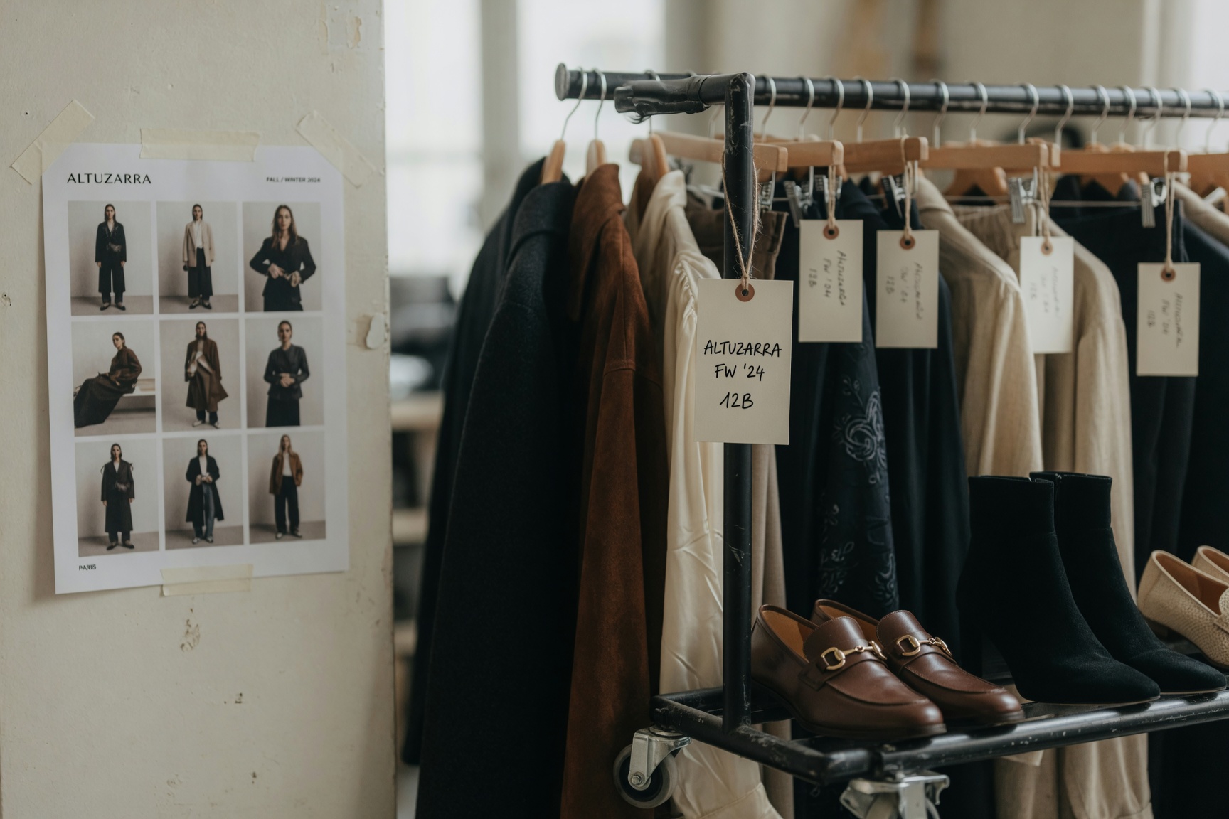 A wardrobe stylist's clothing rack on a photography set, with tagged garments and a look-book reference pinned to the wall.